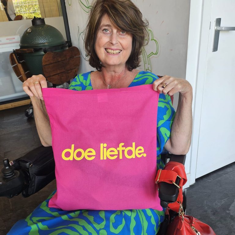 Foto Pien Pien heeft een roze tas in haar handen met "doe liefde" er op gedrukt. Ze heeft een blauw groene jurk aan en kijkt lachend in de camera.
