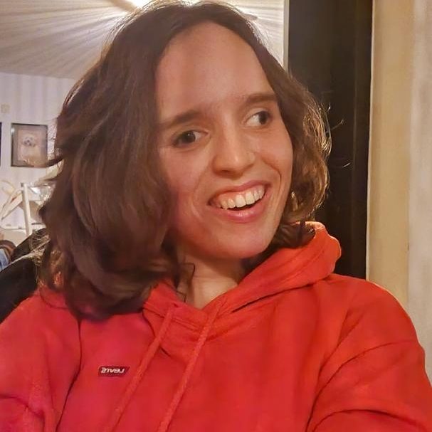Foto Noortje Noortje draagt een rood-oranje trui en lacht naar de camera. Ze heeft half lang bruin haar.
